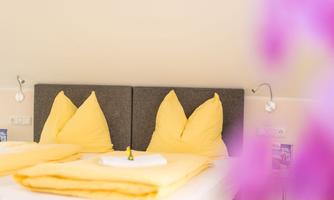 A bright room with a double bed and yellow pillows. A white blanket lies on the bed, and flowers are visible in the background.