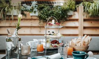 A pretty breakfast table arrangement with various foods, including pastries, fruits, and drinks. In the background, plants are visible, creating a relaxed atmosphere.