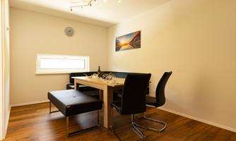 A modern dining room with a table and black chairs. The room has a warm wooden floor and a large window with daylight.