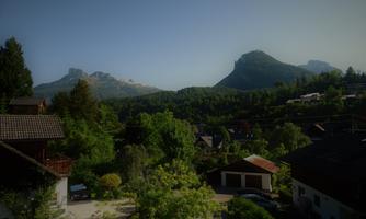 A picturesque landscape with mountains in the background and rustic houses in the foreground. Lush trees and green spaces give the scene a peaceful atmosphere.
