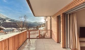 A beautiful balcony with a wooden railing and a view of the mountains. The sunny landscape features snow-covered hills and a clear blue sky.