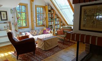 A cozy room with wooden floors and large windows. There is a sofa, bookshelves, and a fireplace that create an inviting atmosphere.