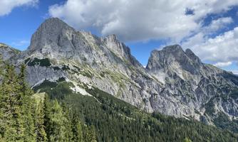 An impressive mountain landscape with steep cliffs and green forests. The sky is partly cloudy and shines through the clouds.