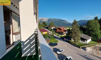 A view from a balcony of a picturesque landscape with mountains and trees. Nearby are residential houses and a quiet street visible.