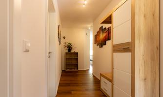 A bright hallway with wooden floors and white walls. On the left side, there is a cabinet and a plant arrangement.