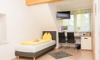 A bright room with a single bed and two chairs at a desk. A small kitchenette and a television are also part of the furnishings.