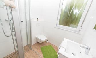 A modern bathroom with a shower, sink, and toilet. The floor is covered with brown tiles, and a green rug lies in front of the sink.