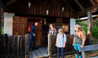 A group of hikers stands in front of a small wooden cabin. They look relaxed and are ready to chat.