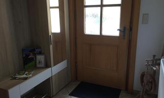 A bright entrance area with a wooden front door and a modern cabinet. A black carpet is on the floor, and there are some decorative items on the shelf.