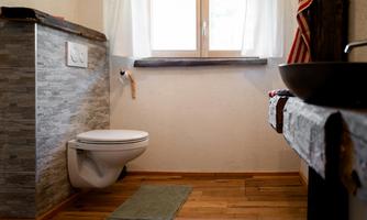 A modern bathroom with a white toilet and a wooden floor. There is a window with bright curtains and a wooden sink shelf.