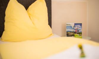 A comfortable bed with yellow pillows and a light duvet. In the background, there are informational brochures about the surrounding area.