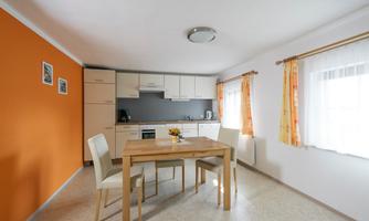 A modern kitchen with a dining table and light chairs. The walls are in neutral colors, with a striking orange accent.