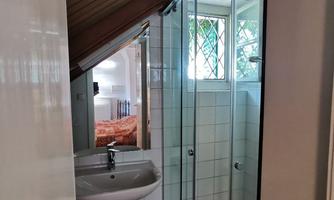 A small bathroom with a sloped roof, equipped with a shower and a sink. The window lets in daylight and you can see part of the bedroom in the background.