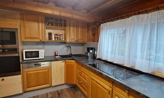 A modern kitchen with wooden details and a large window front. The countertop is made of a dark material, and there are various electrical appliances available.