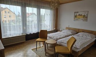 A bright bedroom with two single beds and a small seating area. Large windows provide plenty of daylight.