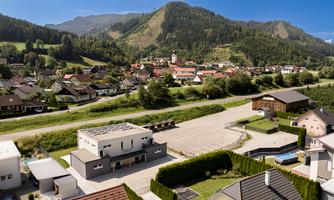 A picturesque landscape with a small village surrounded by mountains. In the foreground, modern houses and a quiet street can be seen.