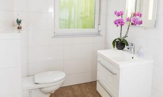 A modern bathroom with white tiles and a large window. It includes a toilet, a sink with storage space, and a yellow bath mat.