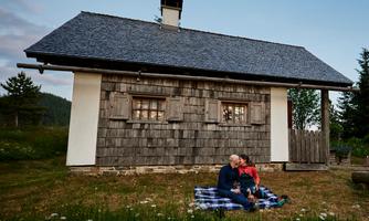 A romantic couple is sitting on a blanket in front of a cozy cabin. The surroundings are green with meadow flowers and gentle hills in the background.