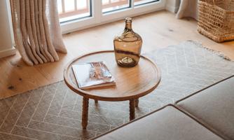 A round wooden table with a decorative glass bottle and a magazine on it. In the background, large windows and bright light can be seen.