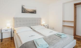 A modern bedroom with a large, bright bed and soft colors. Next to the bed are nightstands with lamps and a spacious wardrobe is visible in the background.