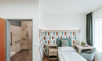 A modern room with a single bed, a cozy armchair, and a small decoration on the table. The adjoining bathroom is bright and designed to be barrier-free.