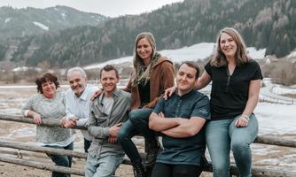 A group of six people stands smiling by a fence in front of a picturesque mountain landscape. It is a friendly and welcoming atmosphere.