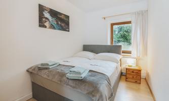 A cozy bedroom with a double bed and soft colors.  
Next to the bed is a nightstand with a lamp and a window overlooking greenery.