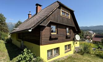 A charming wooden house with a yellow facade and a sloping roof. In the background, mountains and a peaceful landscape can be seen.