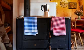 A rustic kitchen with a black stove and two dish towels in blue and red. In the background, there is a wood-paneled room with a dining table.