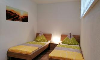 A bright bedroom with two single beds and colorful cushions. On the wall hangs a picture of a landscape with a sunset.