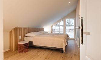 A modern bedroom with a double bed and wooden floor. Large windows offer a view outside and provide plenty of daylight.