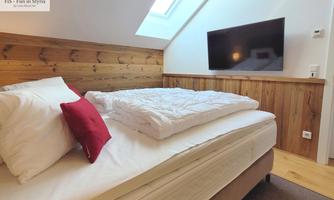 A cozy bedroom with a large bed and red pillows. A TV hangs on the wall, and there is plenty of daylight from the skylight.