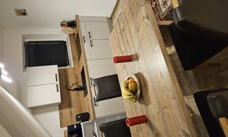 A modern kitchen with a large wooden table and black chairs. On the table, there are fruits and two red candles.