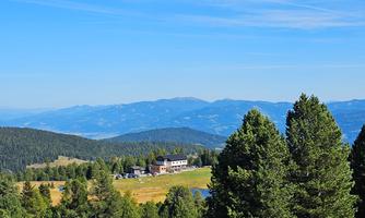A picturesque mountain landscape with green forests and colorful meadows. In the background, the mountains and a clear blue sky are visible.