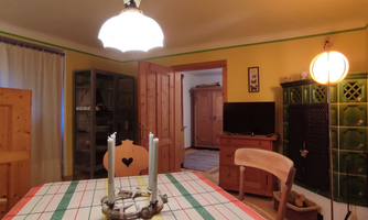 A cozy dining area with a wooden table and a vintage tablecloth. In the background, a wall unit and a television can be seen.