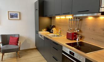 A modern kitchen with black cabinets and wooden surfaces. Next to the kitchen is a comfortable armchair with a red cushion.