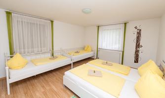 A simple room with two single beds and yellow bed linen. The walls are light and the room has large windows with curtains.