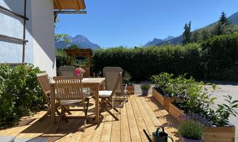 A cozy wooden terrace with a dining table and chairs. Blooming plants and a clear blue sky in the background.