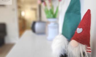 Two cute gnomes wearing hats in red and green stand on a white surface. In the background, fresh flowers in a vase are visible.