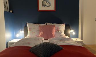 A cozy bedroom with a large bed and colorful pillows. The walls are dark blue, and there are some decorative pictures.