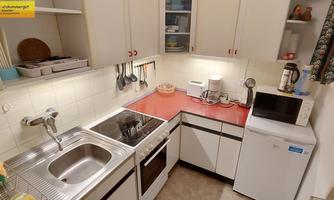 A small, well-equipped kitchen with white cabinets and a red countertop. It includes a stove, a sink, a microwave, and a refrigerator.