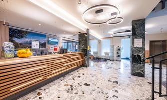 A modern reception area with a wood-paneled reception desk and elegant lighting.  
The floor is made of marbled tiles and large windows let in plenty of daylight.