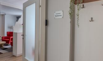 A bright hallway with a closed door leading into a room. In the background, a red sofa and decorative wall moldings can be seen.