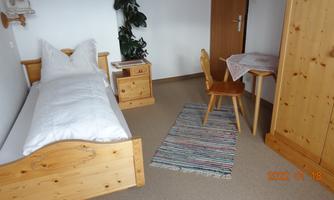 A simple room with a single bed, a nightstand, and a table with a chair. A plant and a rug create a cozy atmosphere.