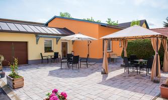 A sunny terrace with garden furniture and umbrellas. The house is painted orange and the area is decorated with plants.