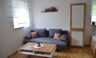 A cozy living room with a gray sofa and a wooden coffee table. The room has large windows that let in plenty of daylight.