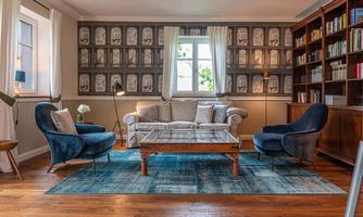 A cozy living room with a bright sofa and blue armchairs. Shelves with books and decorative wallpaper create an inviting atmosphere.