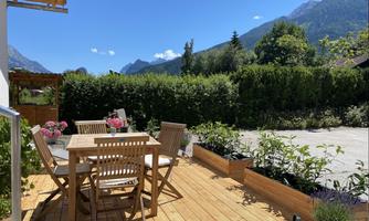 A sunny terrace with wooden furniture and flower beds. In the background, mountains and a green hedge can be seen.