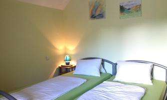 A cozy bedroom with two single beds and light green walls. Two pictures are hanging on the wall, and a lamp provides soft light.