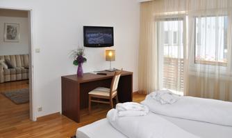 A modern hotel room with two beds, a desk, and a television on the wall. Large windows provide plenty of natural light.